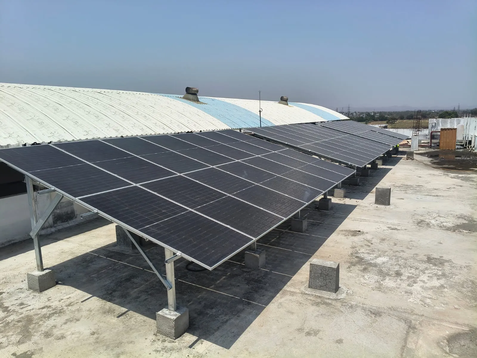 Rows of solar panels on a steel mounting structure on a flat roof beside an arched metal-roof shed.